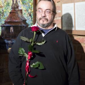 Mężczyzna z wąsami i brodą w okularach w czarnym swetrze, trzyma czerwoną różę. Autor obrazów Paweł Martynów.
