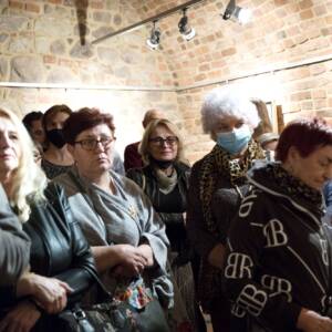 Grupa kobiet patrząca przed siebie w stronę przemawiających osób. Stoją w murowanej, ceglanej piwnicy z zaokrąglonym sufitem.