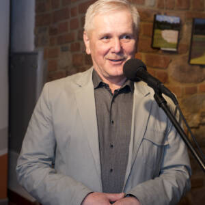 Starszy mężczyzna z siwymi włosami w szarej koszuli i jasnym garniturze przemawia do mikrofonu na statywie.