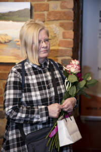Kobieta, blondynka z asymetryczną grzywką w koszuli w dużą czarnobiałą kratę. W rękach trzyma białą różę i małą torebeczkę prezentową.