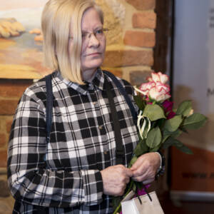 Kobieta, blondynka z asymetryczną grzywką w koszuli w dużą czarnobiałą kratę. W rękach trzyma białą różę i małą torebeczkę prezentową.