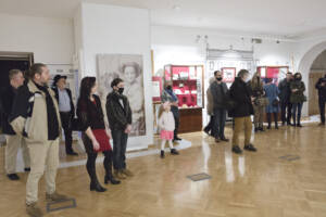 W sali wystawowej stoją osoby w różnym wieku. w centrum stoi dziewczynka w białym sweterku i różowej opasce. W tke gablota z eksponatami.