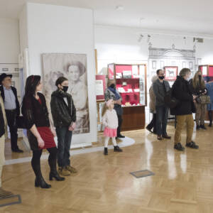 W sali wystawowej stoją osoby w różnym wieku. w centrum stoi dziewczynka w białym sweterku i różowej opasce. W tke gablota z eksponatami.