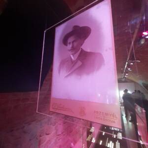 Pod sufitem podwieszona przeźroczysta plexi z nadrukowanym wizerunkiem Janem Szczepanikiem - mężczyzna ze spiczastym wąsem w kapeluszu z okrągłym rondem.