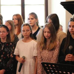 Grupa dziewcząt stojąca równo w środku dziewczynka w białej sukience trzyma mikrofon.