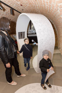 Sala w muzeum z ceglanymi ścianami. Widać dorosłą kobietę zwiedzającą ekspozycję z dwójką dzieci. W centralnym punkcie widać współczesny, biały element ekspozycyjny o łukowatym kształcie.
