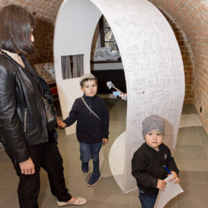 Sala w muzeum z ceglanymi ścianami. Widać dorosłą kobietę zwiedzającą ekspozycję z dwójką dzieci. W centralnym punkcie widać współczesny, biały element ekspozycyjny o łukowatym kształcie.