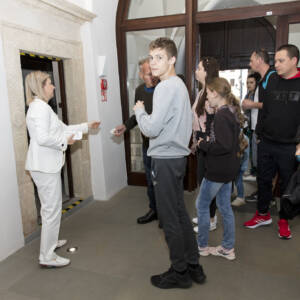 Korytarz-hall w muzeum. Grupa kilku osób czeka przed wejściem na ekspozycję. Osoby wprowadzane są przez młodą kobietę będącą pracownikiem muzeum, ubraną w białe współczesne ubranie.