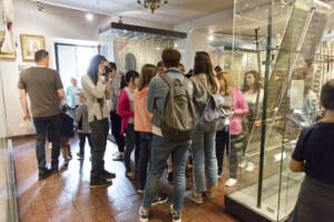 Sala w muzeum. W centralnym puncie grupa kilkunastu osób – głównie młodzież – zwiedzających. W Sali znajdują się gabloty, w których prezentowane są elementy strojów historycznych.