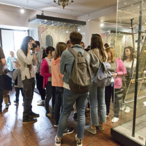 Sala w muzeum. W centralnym puncie grupa kilkunastu osób – głównie młodzież – zwiedzających. W Sali znajdują się gabloty, w których prezentowane są elementy strojów historycznych.