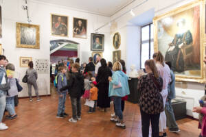 Wysoka sala w muzeum, ze ścianami w kolorze białym. Na ścianach obrazy przedstawiające osoby w strojach historycznych. W Sali grupa osób zwiedzających ekspozycję. Wśród kilkunastu osób zwiedzasjących ekspozycję są osoby dorosłe i dzieci.
