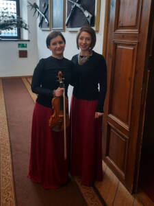 Dwie kobiety w czarnych bluzce i czerwonych spódnicach w ręce trzymają po skrzypcach.