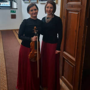 Dwie kobiety w czarnych bluzce i czerwonych spódnicach w ręce trzymają po skrzypcach.
