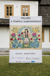 Przed dworem stoi baner z napisem Malarki z powiśla dąbrowskiego. W centrum malowidło kobiety stojącej pośród kwiatów. na dole napis Czerwiec - sierpień 2022