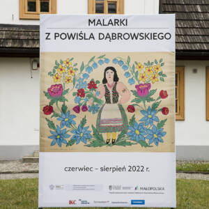 Przed dworem stoi baner z napisem Malarki z powiśla dąbrowskiego. W centrum malowidło kobiety stojącej pośród kwiatów. na dole napis Czerwiec - sierpień 2022