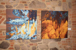 Na ceglanej ścianie wiszą dwa obrazy. Przedstawiają wnętrze jaskini. Oba namalowane są w żywych kolorach. Po lewej dominującym kolorem jest pomarańczowy. Po lewej obraz w dominującym kolorze niebieskim z wystającymi pomarańczowymi pionowymi skalnymi naciekami.