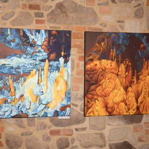 Na ceglanej ścianie wiszą dwa obrazy. Przedstawiają wnętrze jaskini. Oba namalowane są w żywych kolorach. Po lewej dominującym kolorem jest pomarańczowy. Po lewej obraz w dominującym kolorze niebieskim z wystającymi pomarańczowymi pionowymi skalnymi naciekami.