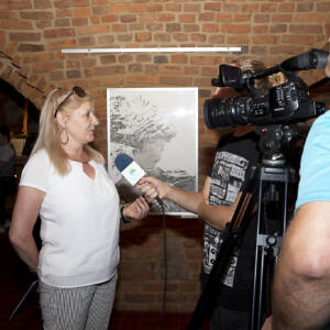 Starsza kobieta z jasnymi włosami udziela wywiadu przed kamerą.