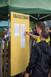 Tablica ogłoszeń z przypiętymi kartkami. Po lewej stronie stoi mężczyzna, sprawdza wyniki biegu.