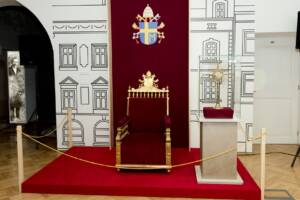 Fotel z czerwoną tapicerką, wysokim oparciem z drewnianymi oparciami i nogami. Na oparciu rzeźbiony herb papieski.