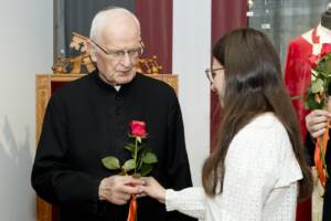 Starszy ksiądz w okularach i siwych włosach przyjmuje czerwoną różę od młodej dziewczyny.