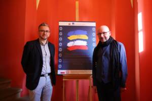 Dwóch mężczyzn w średnim wieku pozuje do zdjęcia stojąc po bokach planszy postawionej na stojaku. Na planszy tytuł wystawy i dwie flagi polska i ukraińska.