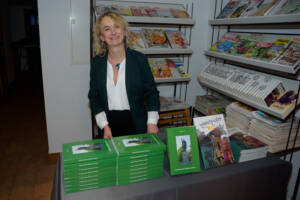 Kobieta za ladą uśmiecha się do zdjęcia. Na ladzie ustawione dwie książki. Jedna zielona niejsza druga większa - komiks.