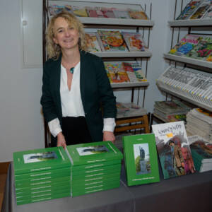 Kobieta za ladą uśmiecha się do zdjęcia. Na ladzie ustawione dwie książki. Jedna zielona niejsza druga większa - komiks.