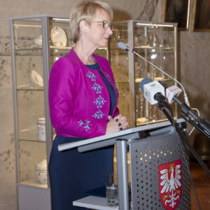 Kobieta z krótkimi blond włosami w okularach przemawia przy mównicy z mikrofonami.