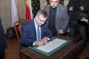Mężczyzna w okularach składa podpis na dokumencie z pieczęciami w zielonej ramie.