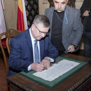 Mężczyzna w okularach składa podpis na dokumencie z pieczęciami w zielonej ramie.