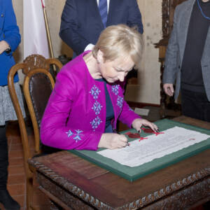 Kobieta, blondynka z krótkimi włosami składa podpis na dokumencie z pieczęciami w zielonej ramie.