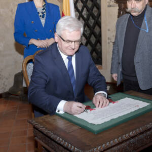 Mężczyzna w okularach i siwymi włosami składa podpis na dokumencie z pieczęciami w zielonej ramie.