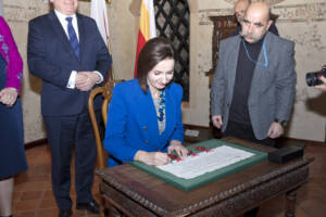 Kobieta, brunetka w niebieskiej garsonce składa podpis na dokumencie z pieczęciami w zielonej ramie.
