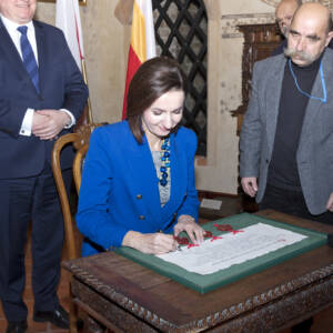 Kobieta, brunetka w niebieskiej garsonce składa podpis na dokumencie z pieczęciami w zielonej ramie.