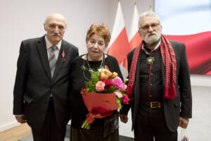 Troje starszych osób pozuje do zdjęcia. Kobieta na środku trzyma kwiaty.