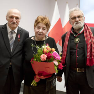 Troje starszych osób pozuje do zdjęcia. Kobieta na środku trzyma kwiaty.