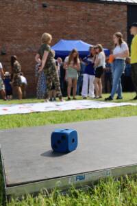 na pierwszym planie duża niebieska kostka do gry. W tle kobieta w spodniach moro omawia szczegóły gry z grupką młodzieży.