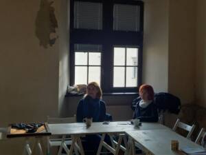 Dwie skupione kobiety siedzące przy stole. Za nimi znajdują się dwa okna.