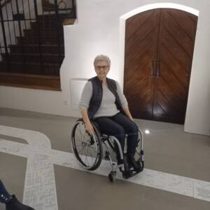 Starsza kobieta z uśmiechem na twarzy siedzi na wózku i trzyma ręce na kołach wózka. W tle drewniane drzwi przypominające portal oraz drewniane schody.