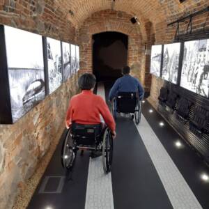 Dwie osoby, kobieta i mężczyzna, poruszający się na wózku wzdłuż korytarza. Na ceglanych ścianach znajdują się zdjęcia multimedialne.